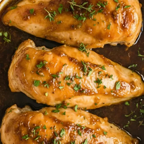 A close-up of Oven Baked Brown Sugar Garlic Chicken breasts in a baking dish, featuring a glossy caramelized glaze and garnished with fresh thyme.