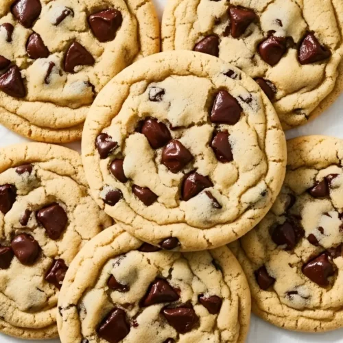 Bakery style chocolate chip cookies stacked on a wooden surface, featuring thick chewy centers, molten chocolate pools, and golden crinkly edges.