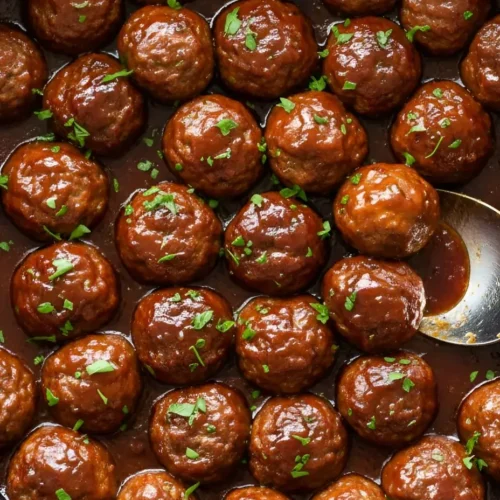 Close-up of glossy grape jelly meatballs simmering in a black slow cooker with a thick BBQ chili sauce glaze and fresh parsley garnish.