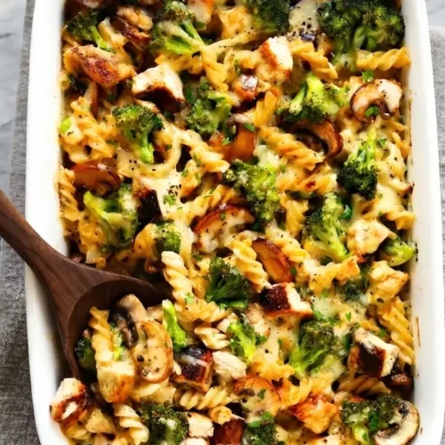 Gemini said A close-up, top-down photograph of a healthy broccoli chicken casserole in a white ceramic baking dish. The casserole features cooked spiral pasta, roasted chicken pieces, broccoli florets, and sliced mushrooms in a creamy sauce, all baked with melted cheese and fresh parsley garnish