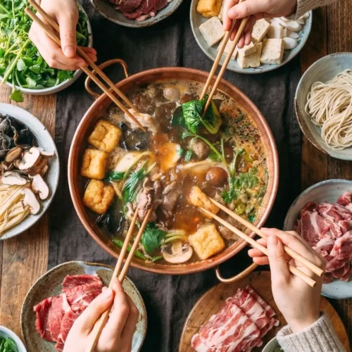 A bubbling split pot of Chinese hot pot at home, surrounded by plates of thinly sliced beef, fresh vegetables, and dipping sauces.
