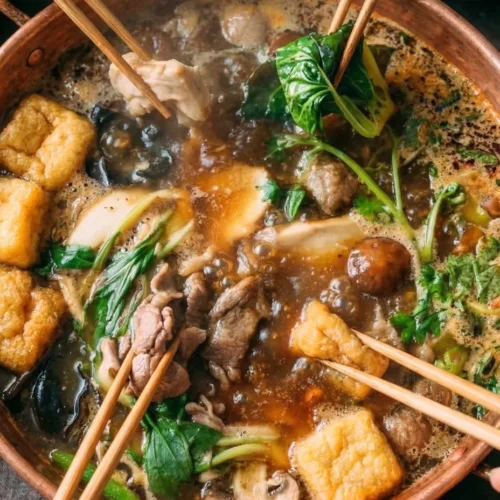 A close-up of a stainless steel split pot sitting securely on a tabletop portable butane burner, ready for a homemade Chinese hot pot dinner with small wire mesh strainers resting on the side.