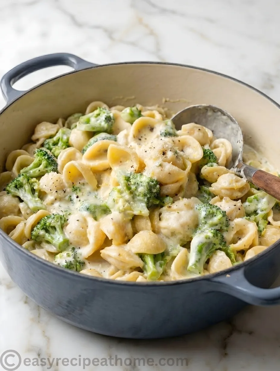 A close-up, overhead shot of creamy broccoli pasta served in a speckled ceramic bowl. Tender, cup-shaped pasta shells and bright green, bite-sized broccoli florets are thickly coated in a glossy, rich cheese sauce.