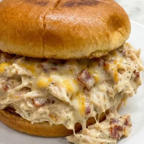 A toasted hamburger bun heavily loaded with thick, shredded cracked chicken crockpot meat covered in a creamy bacon and cheddar cheese sauce.