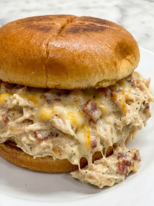 A toasted hamburger bun heavily loaded with thick, shredded cracked chicken crockpot meat covered in a creamy bacon and cheddar cheese sauce.