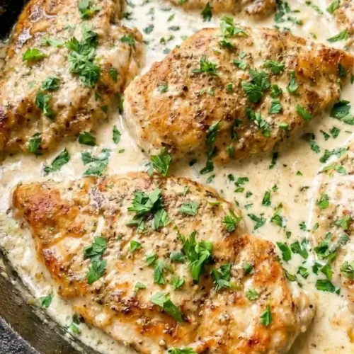 A heavy skillet filled with fiercely seared chicken cutlets heavily dripping in a wildly rich, bright white garlic parmesan cream sauce garnished with green parsley.