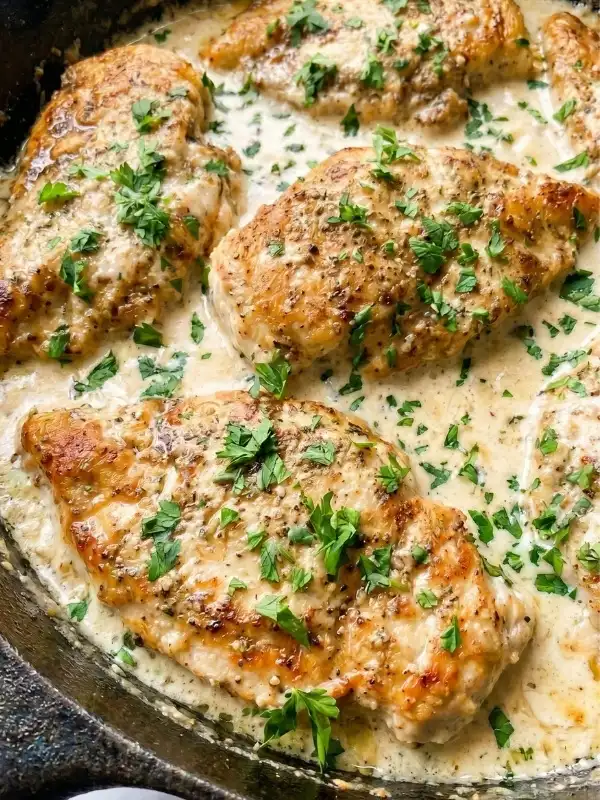 A heavy skillet filled with fiercely seared chicken cutlets heavily dripping in a wildly rich, bright white garlic parmesan cream sauce garnished with green parsley.