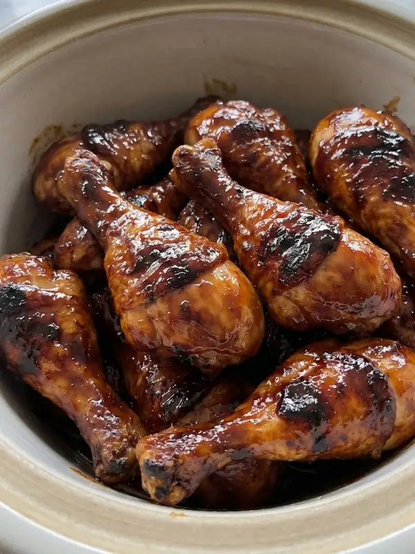 A heavy ceramic slow cooker filled with bone-in crock pot chicken drumsticks heavily coated in a thick, dark molasses barbecue sauce and dry spice rub.