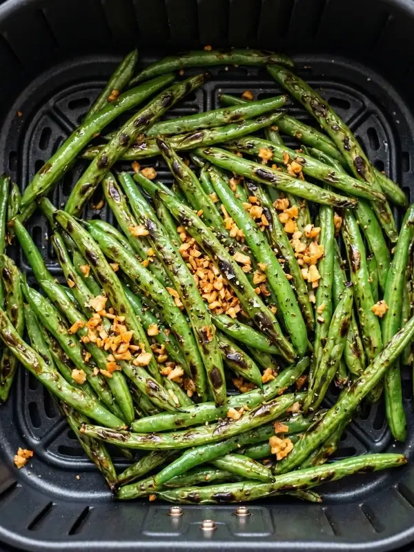 A close-up view of a warm AIR FRYER BOX filled with hot green beans, clearly showing the highly textured, wrinkled skins glistening with olive oil and topped with heavily toasted pieces of raw garlic.