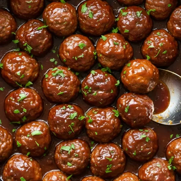 Close-up of glossy grape jelly meatballs simmering in a black slow cooker with a thick BBQ chili sauce glaze and fresh parsley garnish.