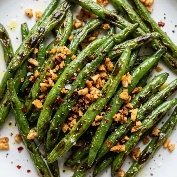 A wide, matte white ceramic dinner plate heavily loaded with vibrant, blistered air fryer green beans, showing deeply charred edges and coated entirely in golden-brown, crispy minced garlic chips and coarse sea salt.